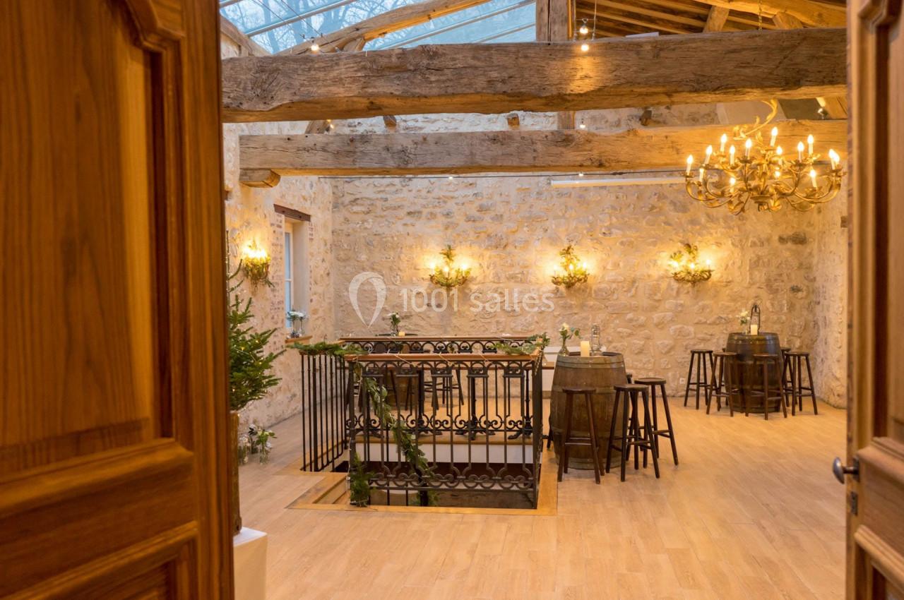 Salle en pierre avec poutres apparentes, lustres, tables hautes et décoration végétale, vue depuis une porte ouverte.