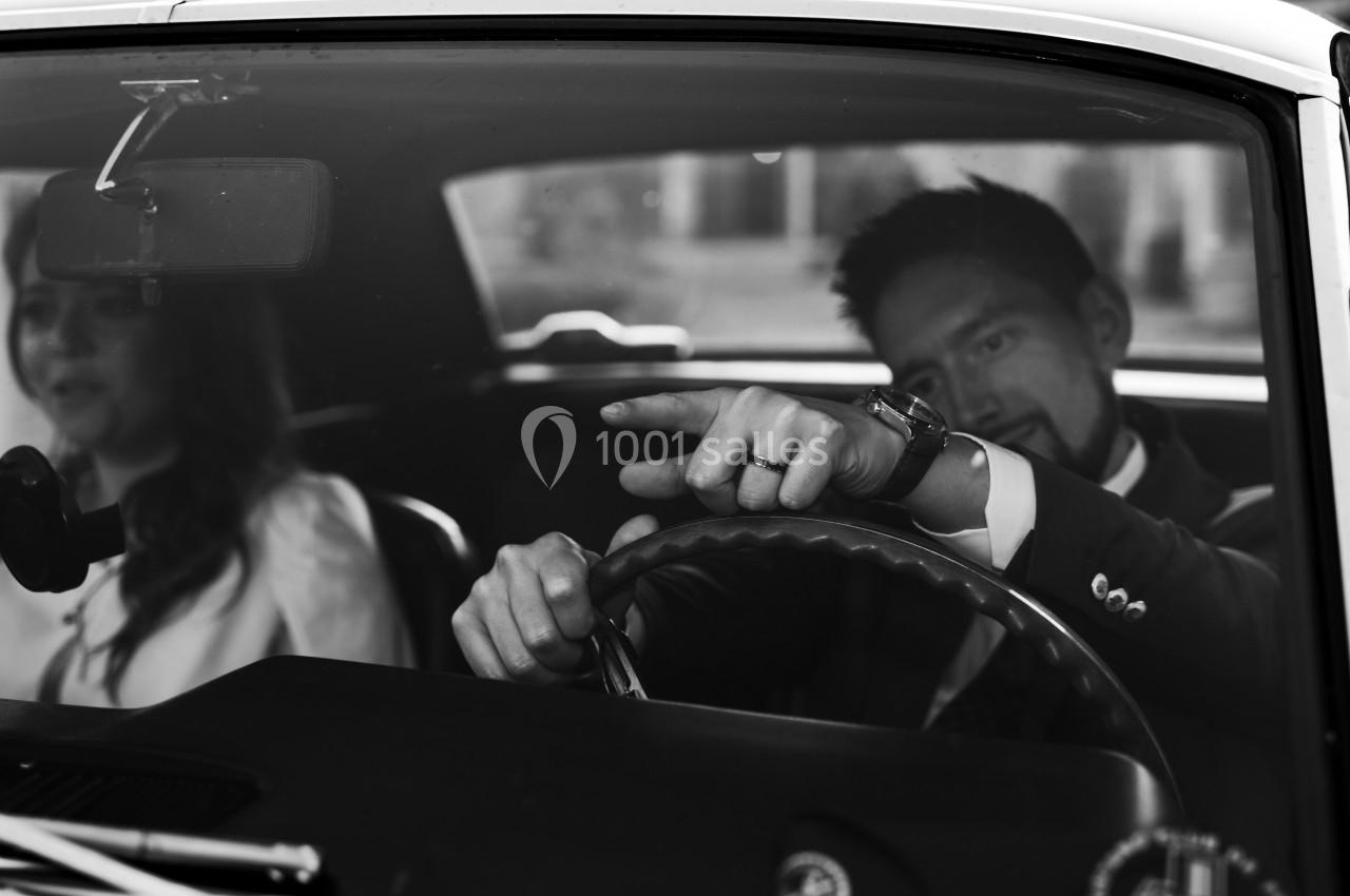 Un homme en costume conduit une voiture ancienne, accompagné d'une femme assise sur le siège passager.