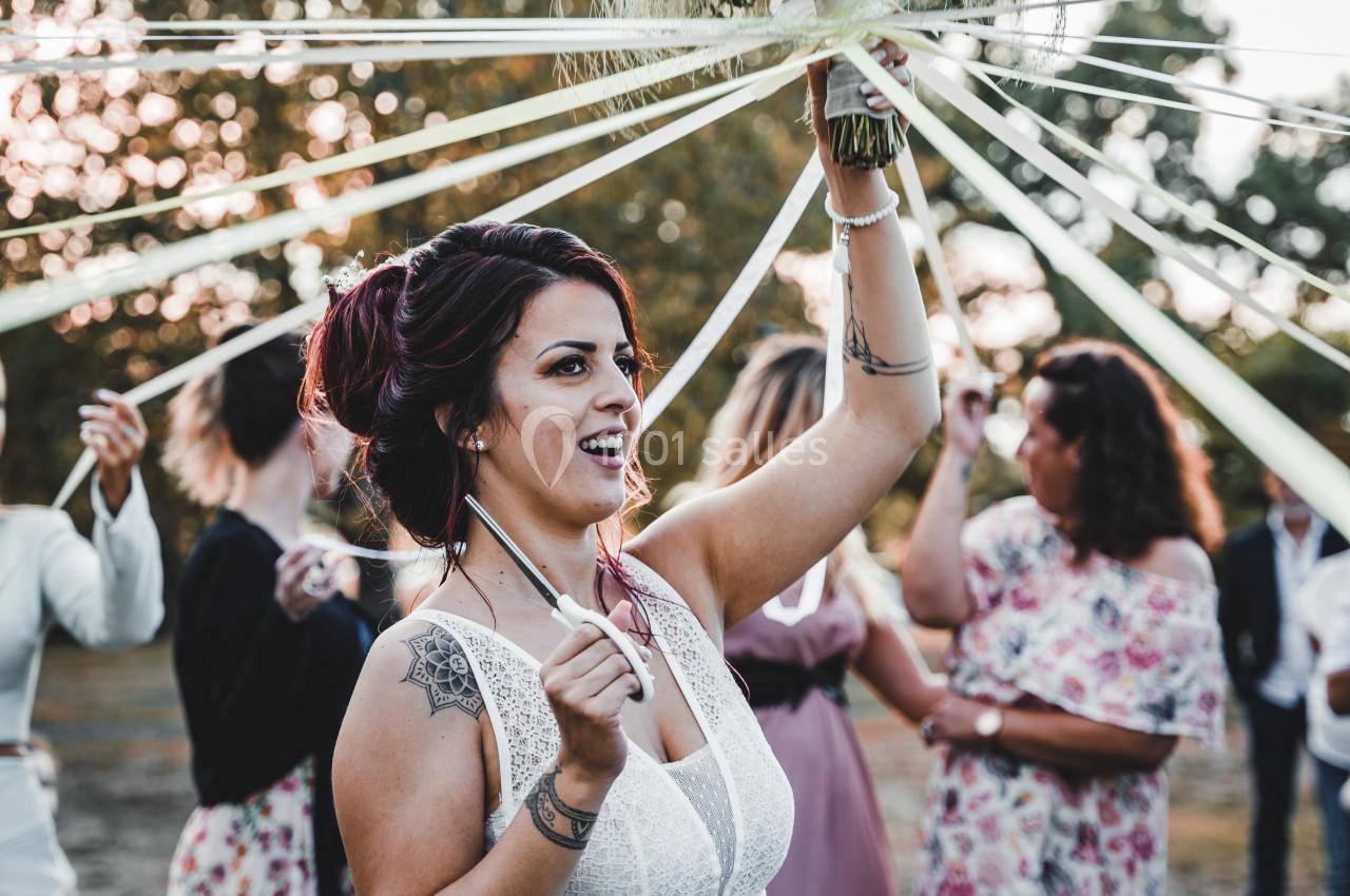 Une femme en robe blanche tient un bouquet entouré de rubans, entourée de personnes lors d'une cérémonie en extérieur.