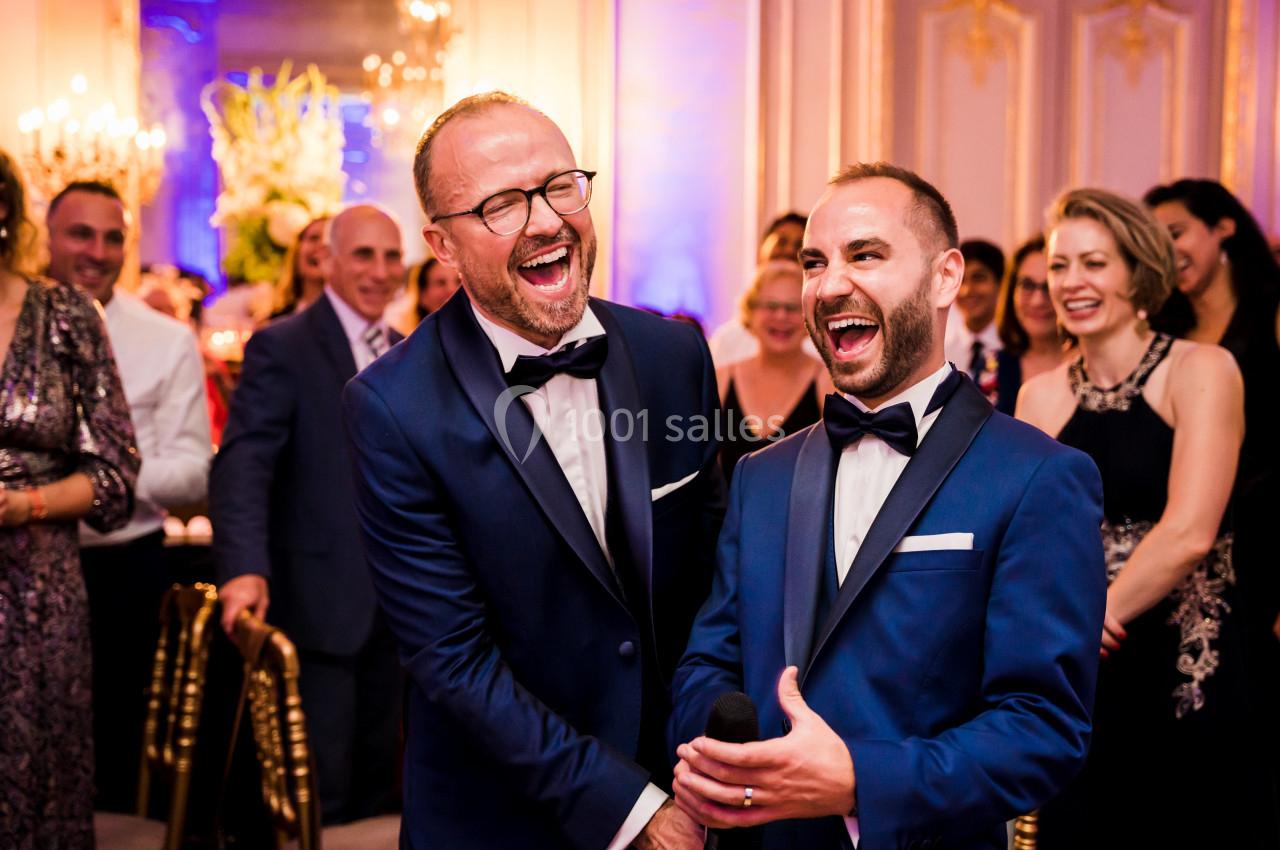 Deux hommes en costume souriant et riant lors d'un événement festif entourés d'invités élégants.