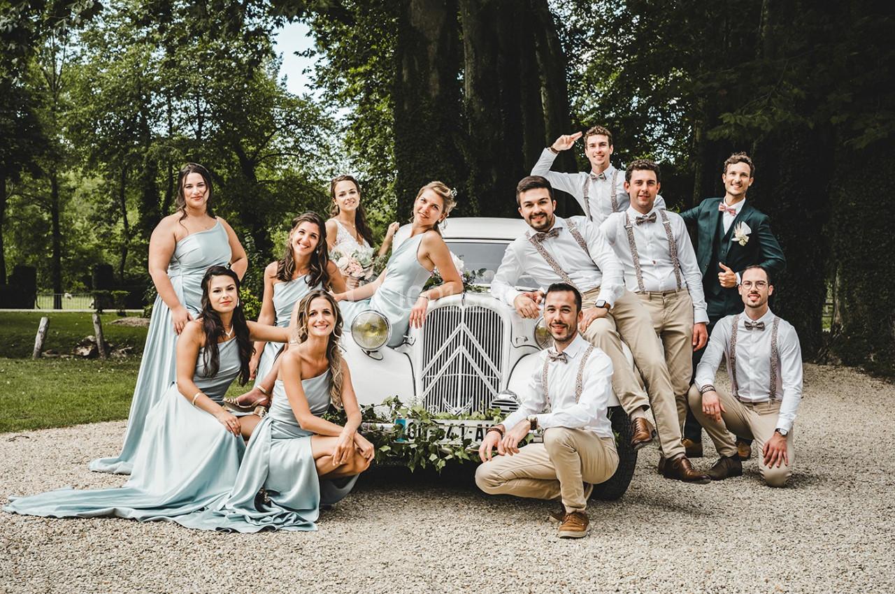 Un groupe de personnes posant autour d'une voiture ancienne blanche dans un cadre extérieur verdoyant.