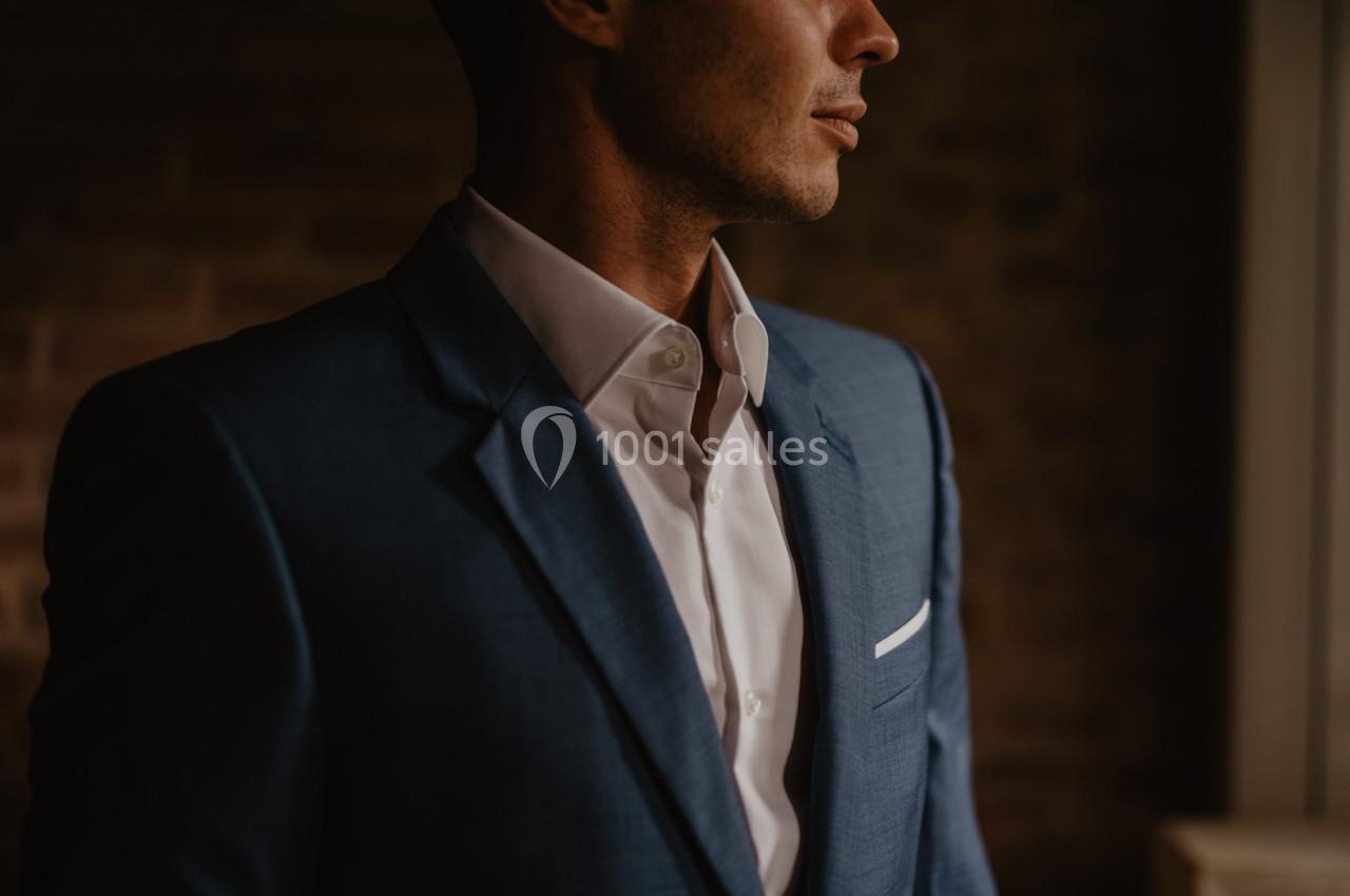 Homme en costume bleu et chemise blanche, partiellement éclairé, devant un mur de briques sombre.