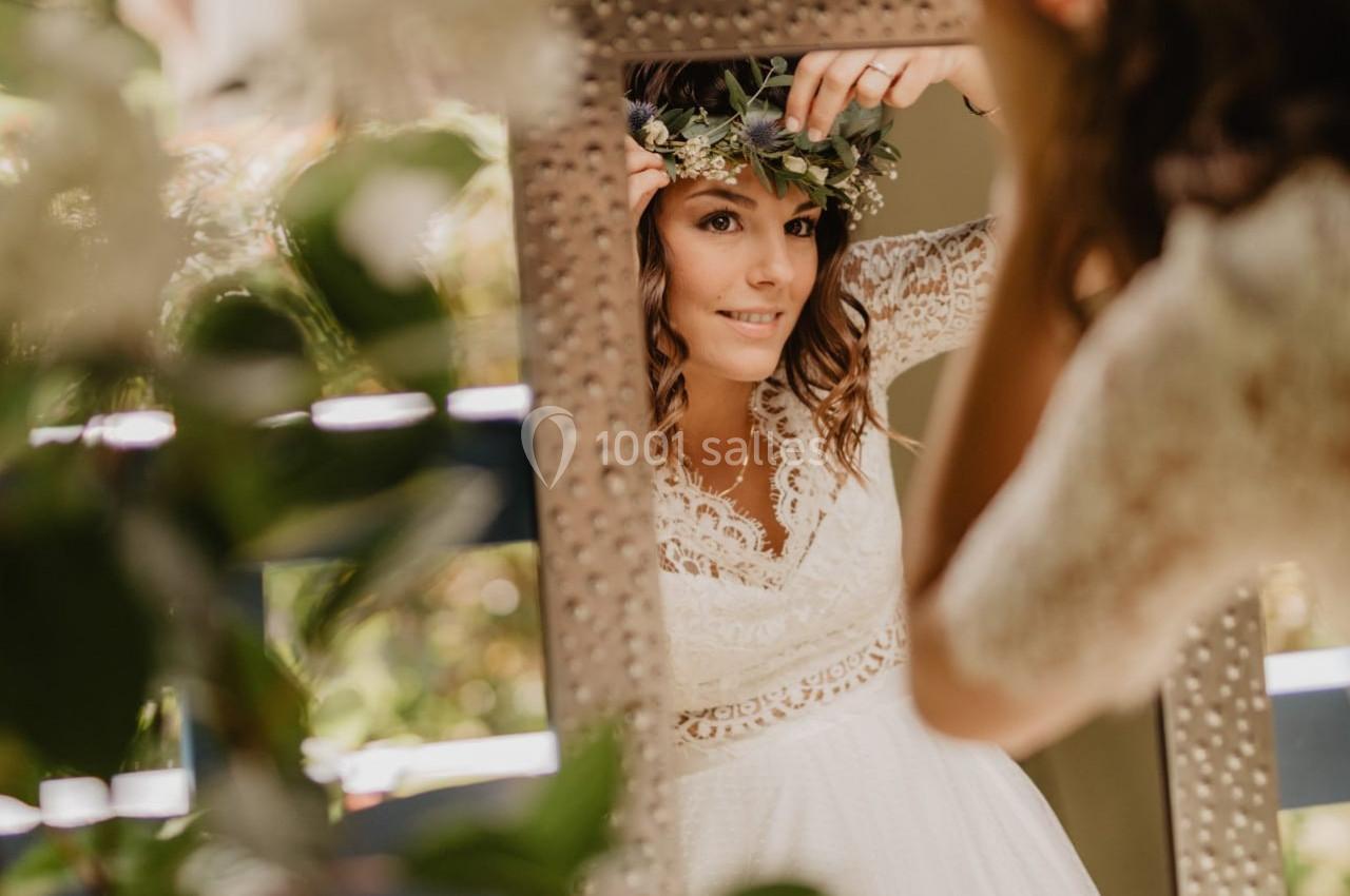 Une femme en robe de mariée ajuste une couronne de fleurs devant un miroir, entourée de verdure.