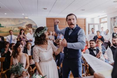 Un couple assis lors d'une cérémonie, l'homme ému essuie une larme tandis que la femme le regarde avec tendresse.