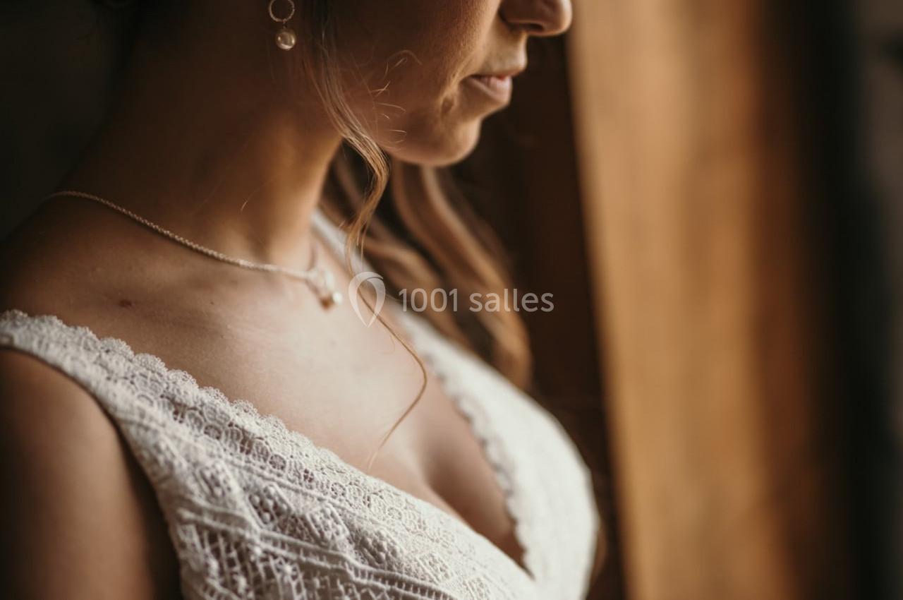 Femme en robe de mariée en dentelle blanche, coiffée d'une couronne florale, regardant par une fenêtre en bois.