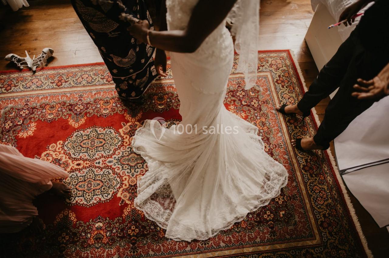 Une mariée en robe blanche est entourée de personnes sur un tapis rouge orné de motifs complexes.