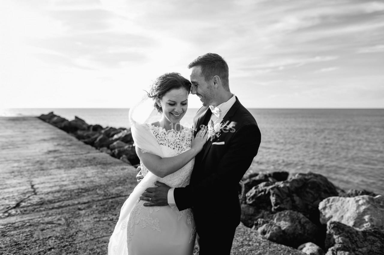 Un couple en tenue de mariage sourit et s'enlace près d'une jetée face à la mer, sous un ciel dégagé.