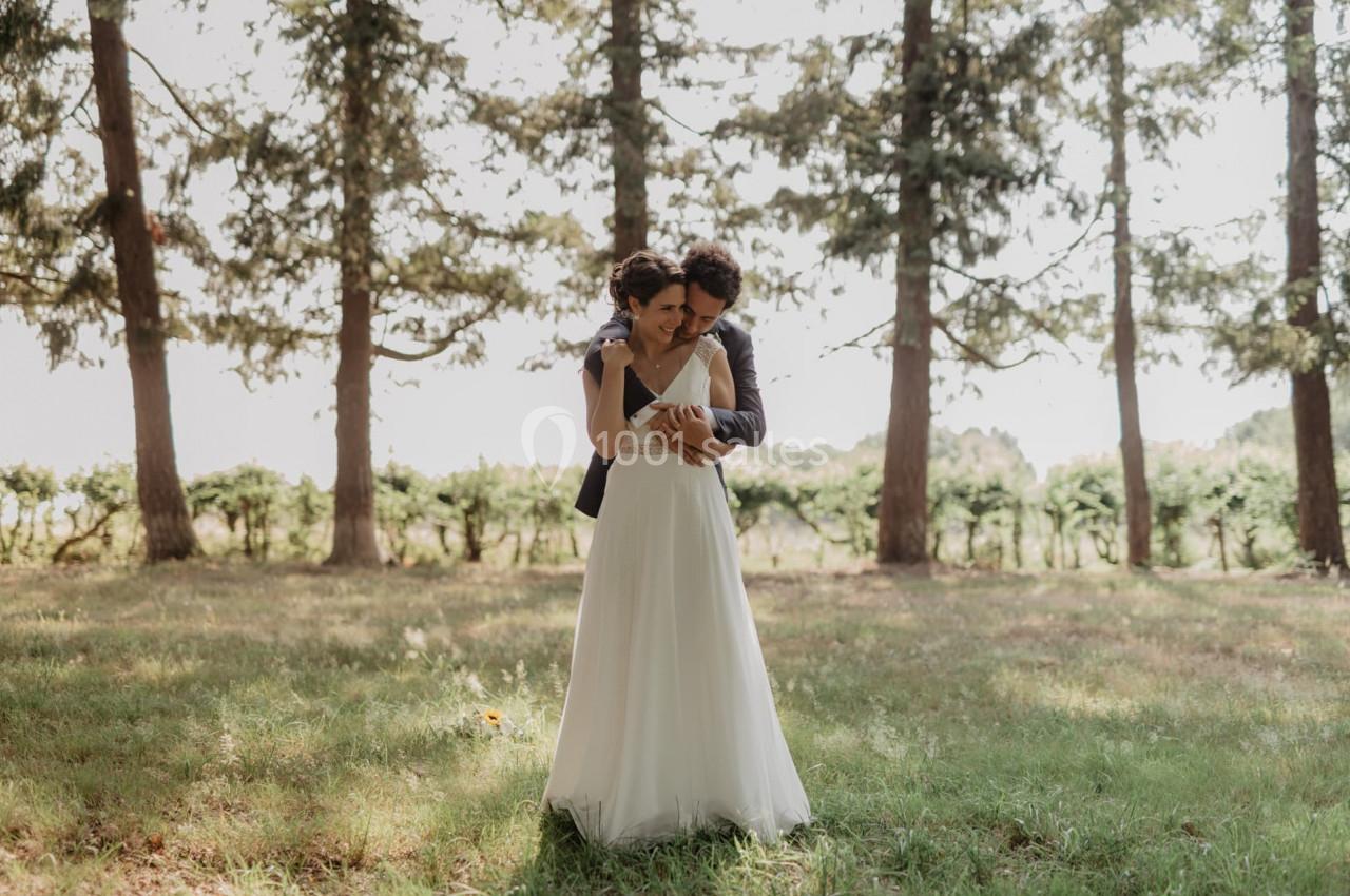 Un couple en tenue de mariage s'enlace dans un champ ensoleillé bordé d'arbres.