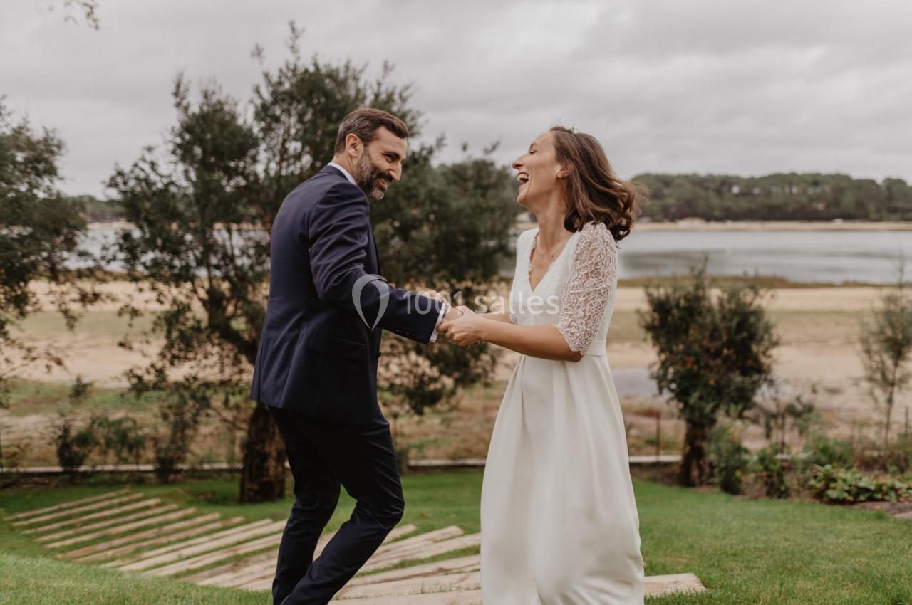 Un couple en tenue de mariage danse joyeusement sur une pelouse avec un paysage naturel en arrière-plan.