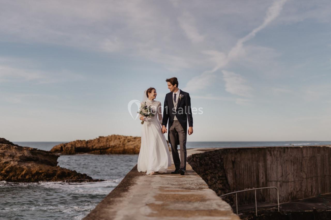 Un couple en tenue de mariage marche main dans la main sur une jetée au bord de la mer, sous un ciel dégagé.