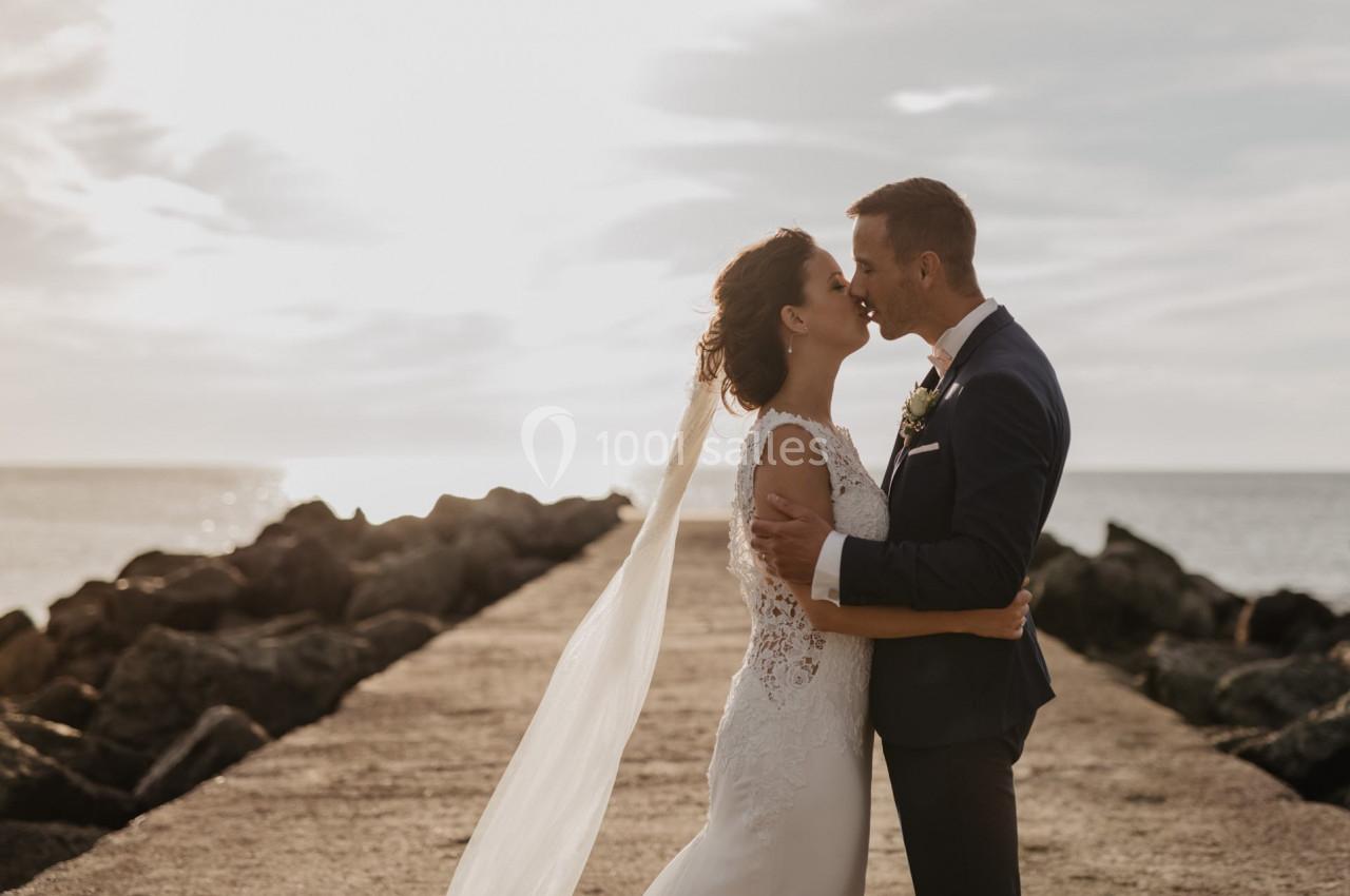 Un couple en tenue de mariage s'embrasse sur une jetée au bord de la mer, sous une lumière douce.