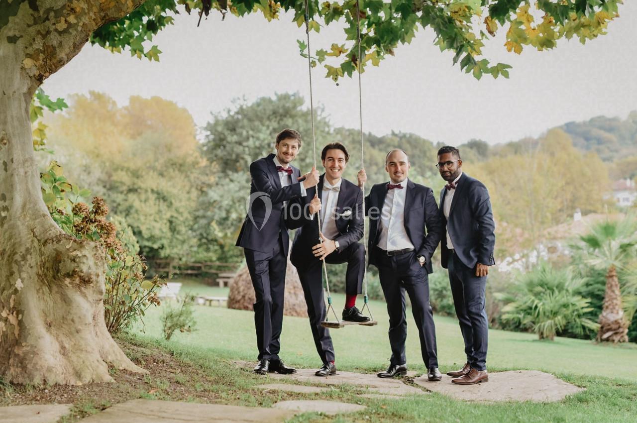 Quatre hommes en costume posent autour d'une balançoire suspendue à un arbre dans un jardin verdoyant.