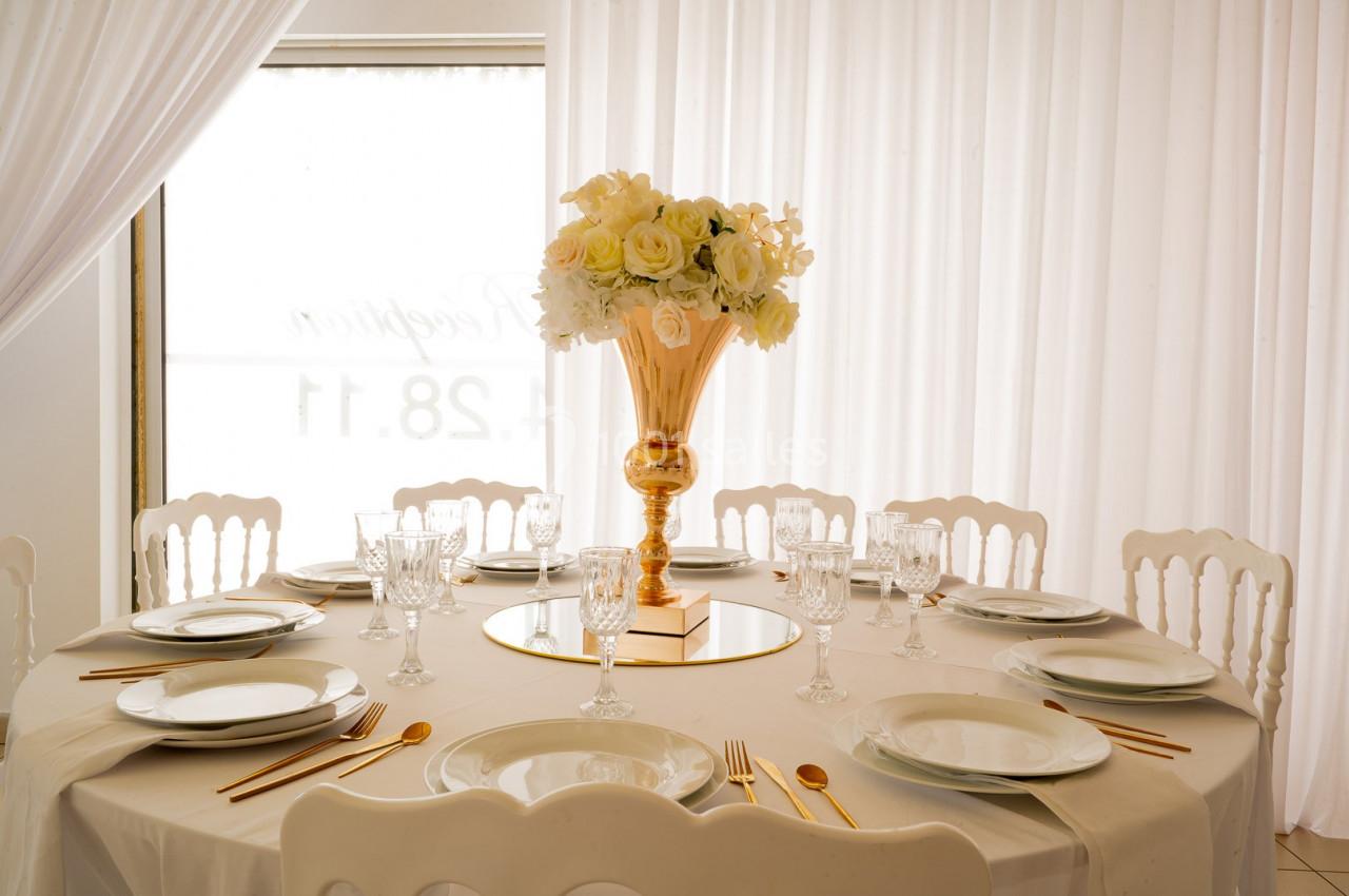 Table ronde élégamment dressée avec une nappe blanche, vaisselle raffinée et un vase doré rempli de fleurs blanches.