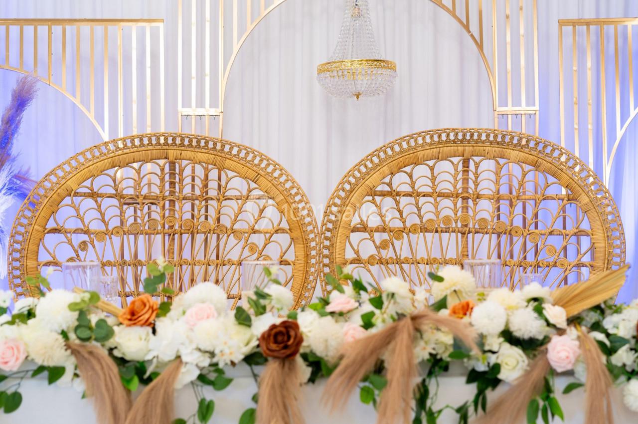 Deux fauteuils en rotin doré devant une table ornée de fleurs et d'herbes séchées, avec un lustre en arrière-plan.