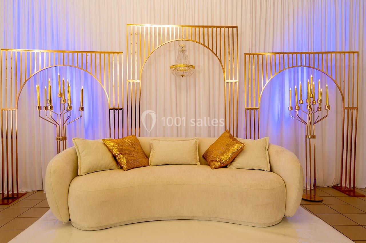 Canapé beige avec coussins dorés, entouré de chandeliers et d'arcs métalliques dorés sur fond de rideaux blancs.