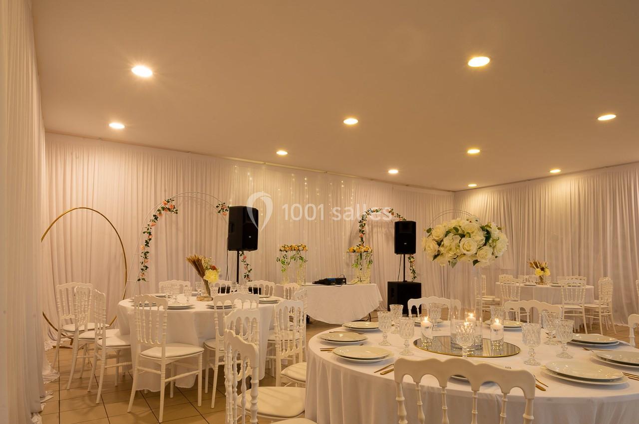 Salle de réception décorée avec des tables rondes, des chaises blanches, des fleurs et des arches lumineuses.
