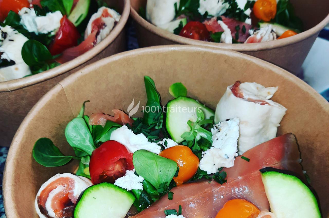 Salade composée dans un bol en carton avec légumes frais, tomates cerises, fromage, jambon cru et herbes.