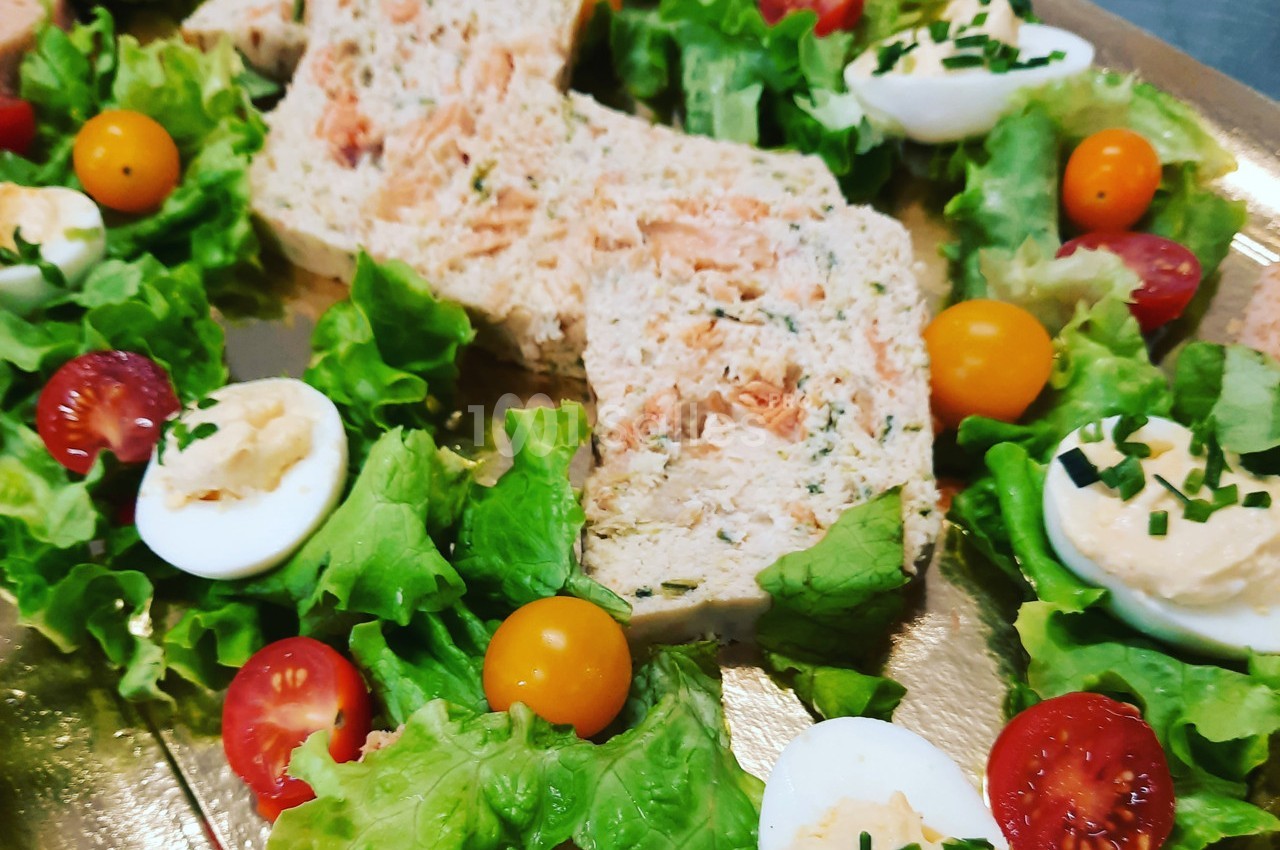 Terrine de poisson entourée de feuilles de salade, tomates cerises, œufs durs garnis et ciboulette sur un plateau doré.