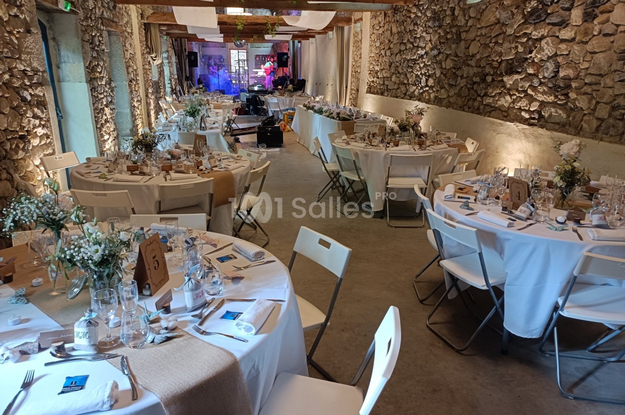 Salle en pierre aménagée pour un événement, avec tables rondes décorées et chaises blanches.