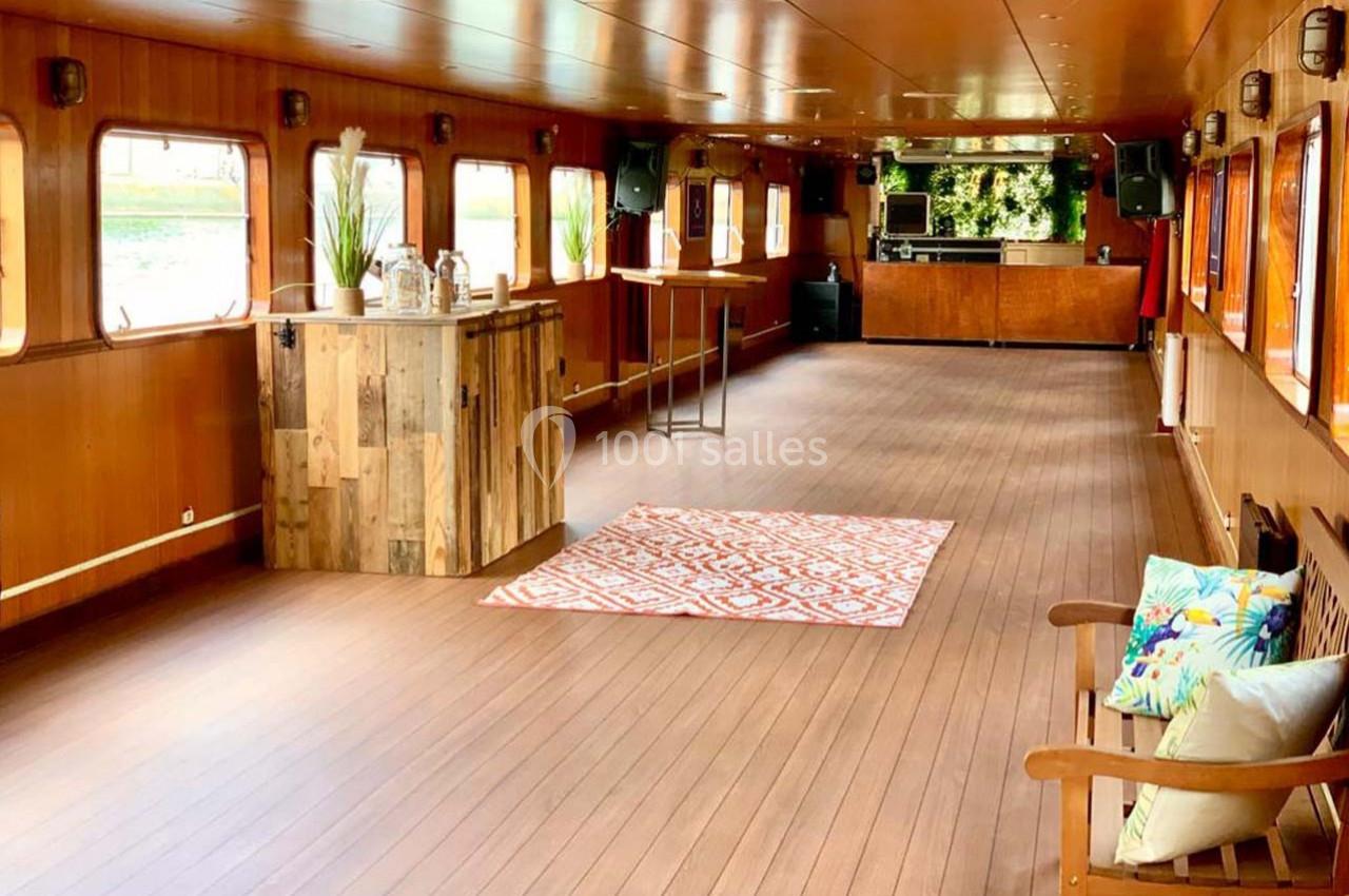 Intérieur d'une péniche aménagée avec un parquet en bois, un bar en bois, des plantes et des sièges décorés de coussins.