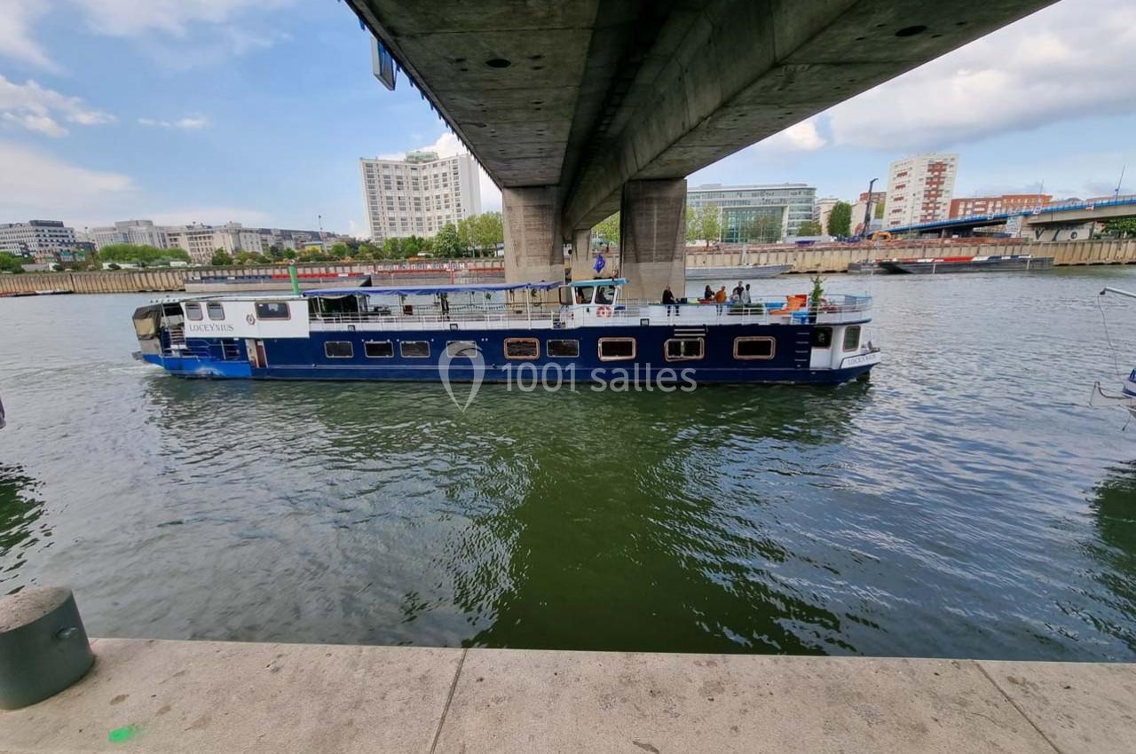 Bateau bleu naviguant sous un pont sur une rivière, entouré de bâtiments urbains et d'une rive aménagée.