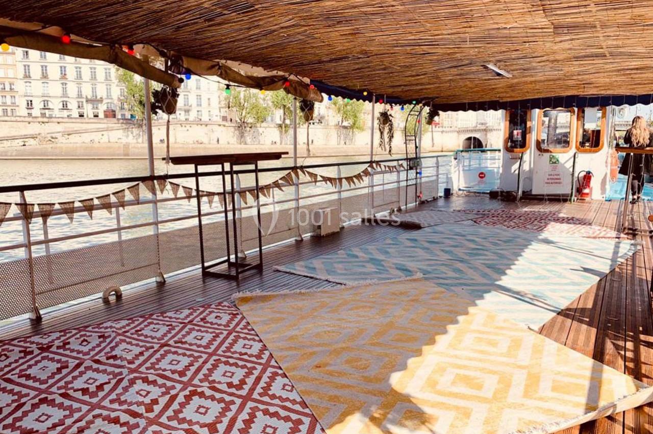 Terrasse d'une péniche avec tapis colorés, toit en bambou et vue sur des bâtiments en bord de rivière.