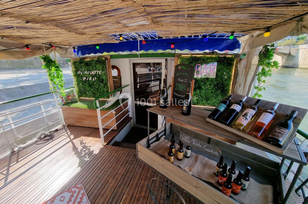 Terrasse en bois d'un bateau avec bar, étagères de bouteilles de vin, décoration végétale et guirlandes lumineuses.