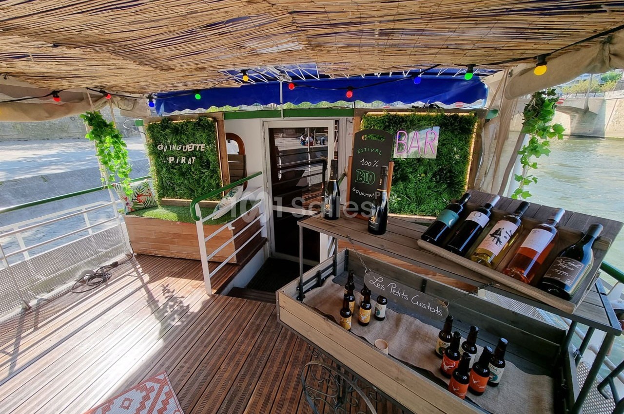 Terrasse en bois d'un bateau avec bar, étagères de bouteilles de vin, décoration végétale et guirlandes lumineuses.