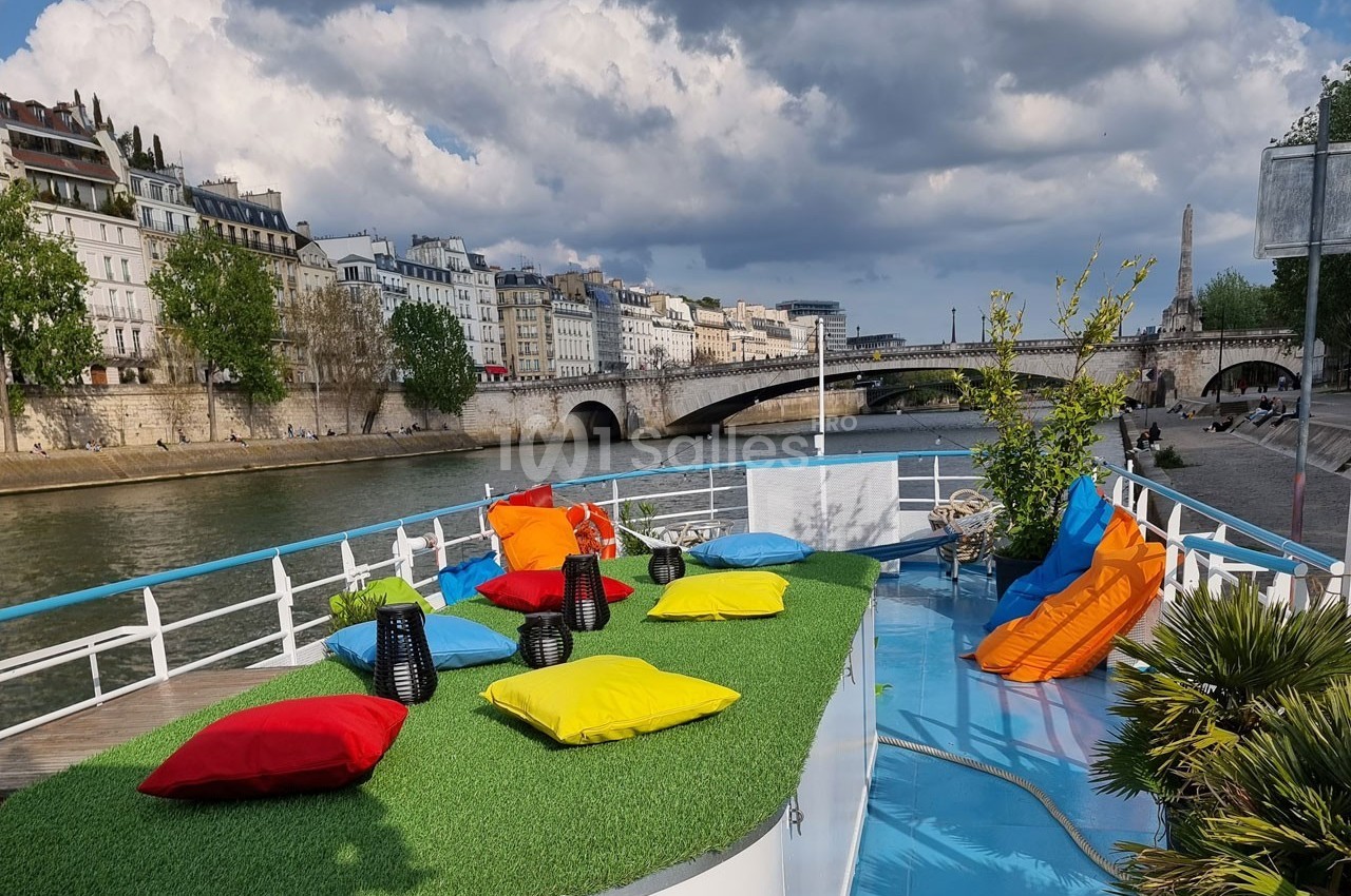 Terrasse colorée avec coussins et plantes sur une péniche, vue sur la Seine et des immeubles parisiens en arrière-plan.