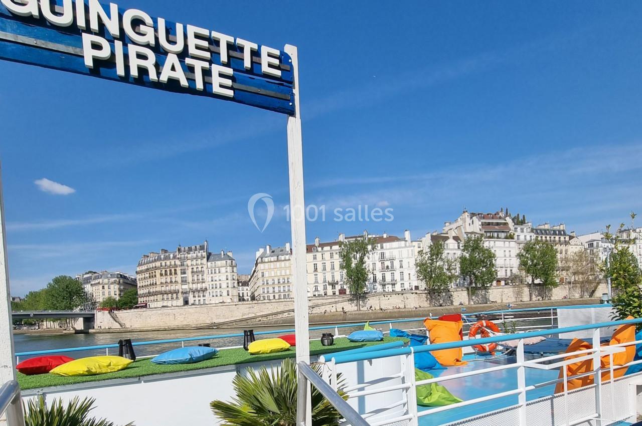 Terrasse colorée d'une péniche avec coussins et vue sur des immeubles historiques au bord d'une rivière.