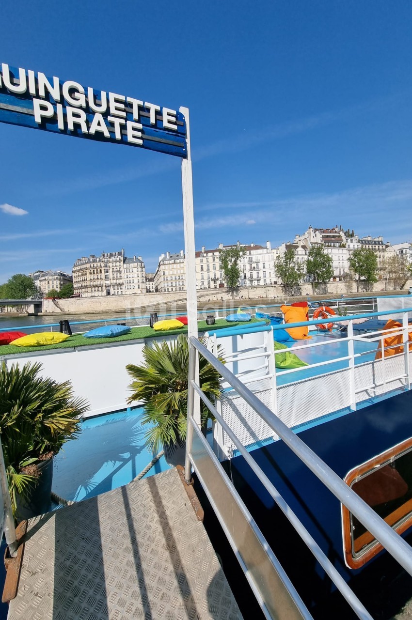 Passerelle menant à une péniche colorée avec vue sur des bâtiments historiques en bord de rivière.