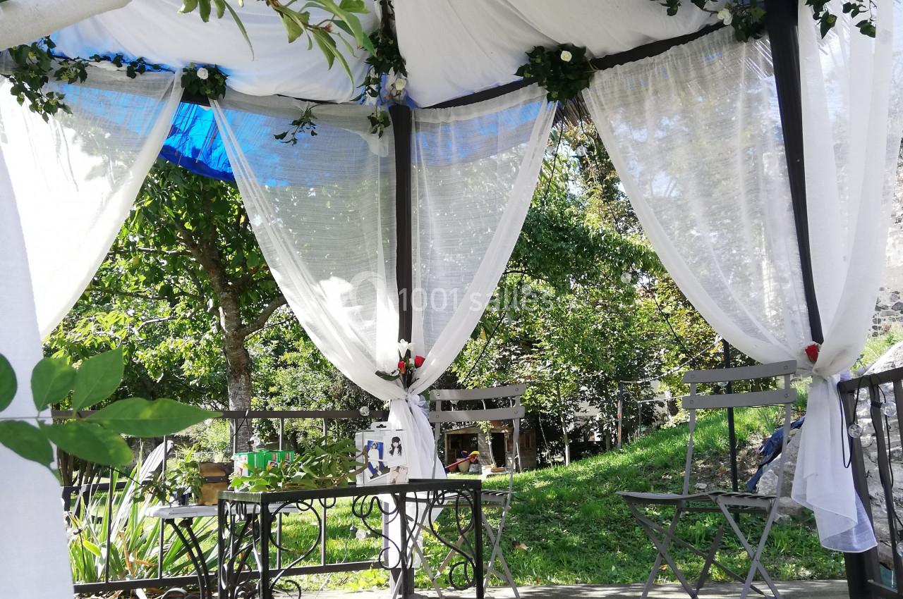 Tonnelle décorée de voilages blancs et de feuillage, avec table et chaises sur une terrasse ensoleillée.