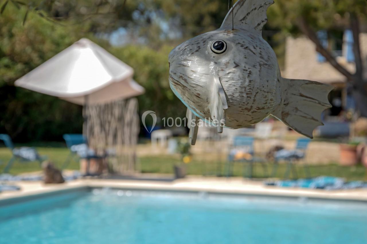 Décoration en forme de poisson suspendue près d'une piscine dans un jardin avec des chaises et un parasol en arrière-plan.
