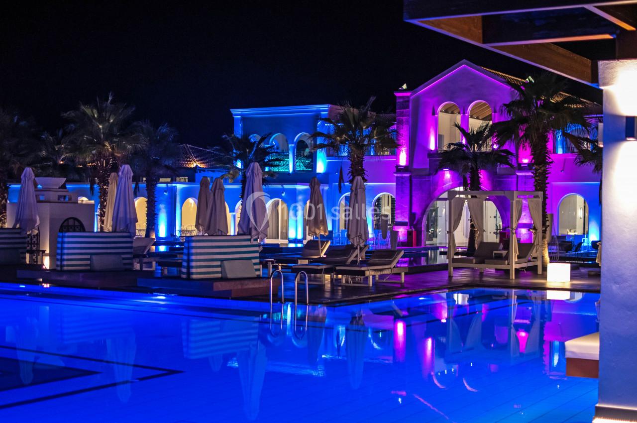 Piscine éclairée de nuit entourée de transats, parasols fermés et bâtiments illuminés aux tons colorés.