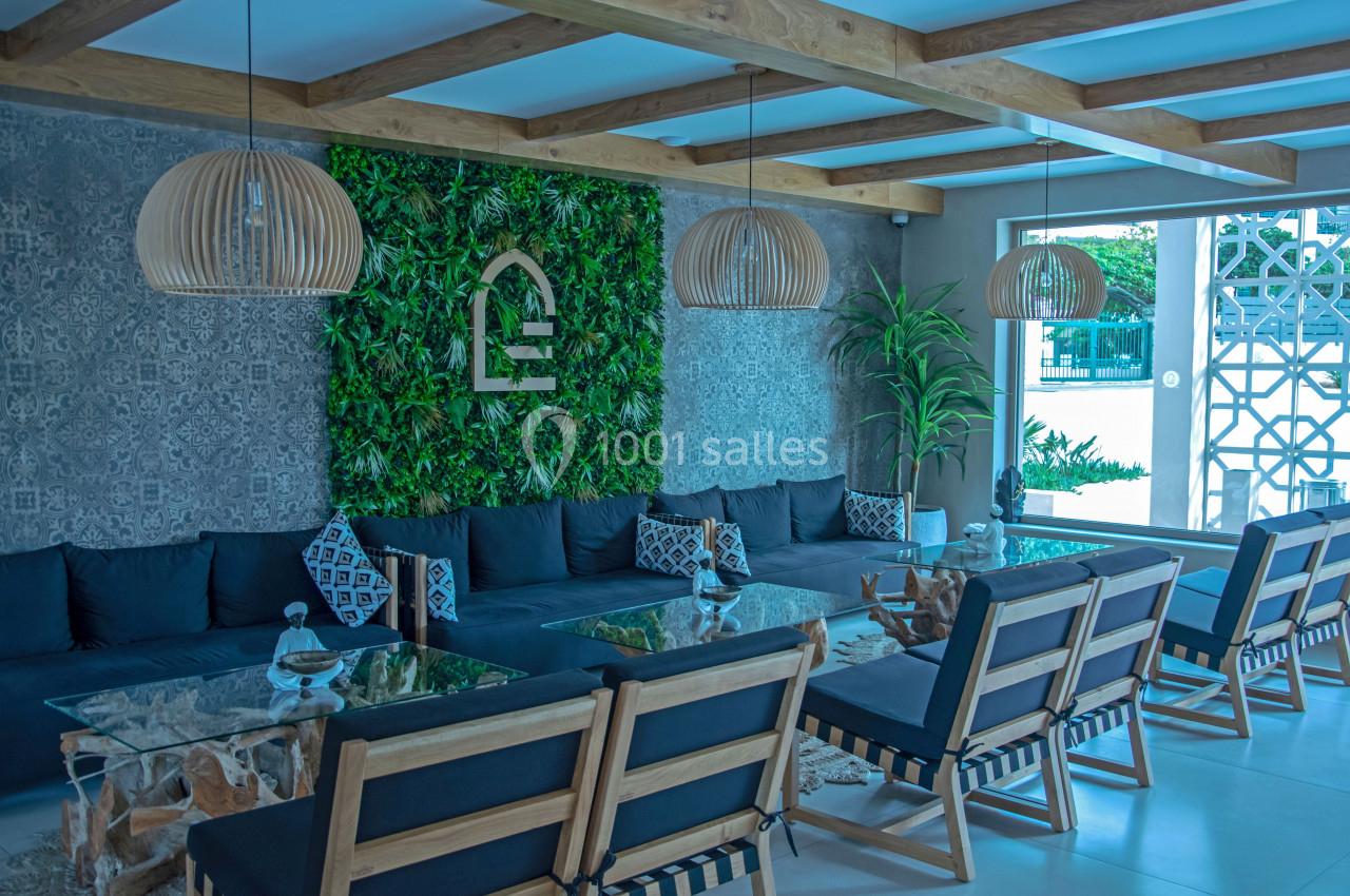 Salon moderne avec murs végétalisés, canapés noirs, tables en verre et chaises en bois sous des luminaires suspendus.