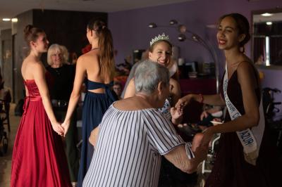 Des jeunes femmes en robes de soirée interagissent avec des résidents d'un établissement, dans une ambiance conviviale.