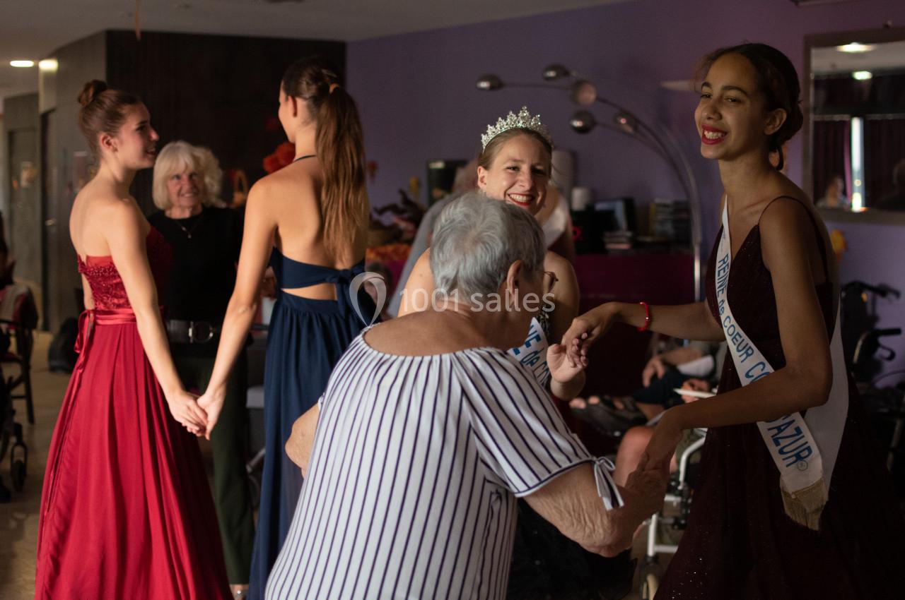 Des jeunes femmes en robes de soirée interagissent avec des résidents d'un établissement, dans une ambiance conviviale.