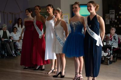 Des jeunes femmes en robes de soirée interagissent avec des résidents d'un établissement, dans une ambiance conviviale.