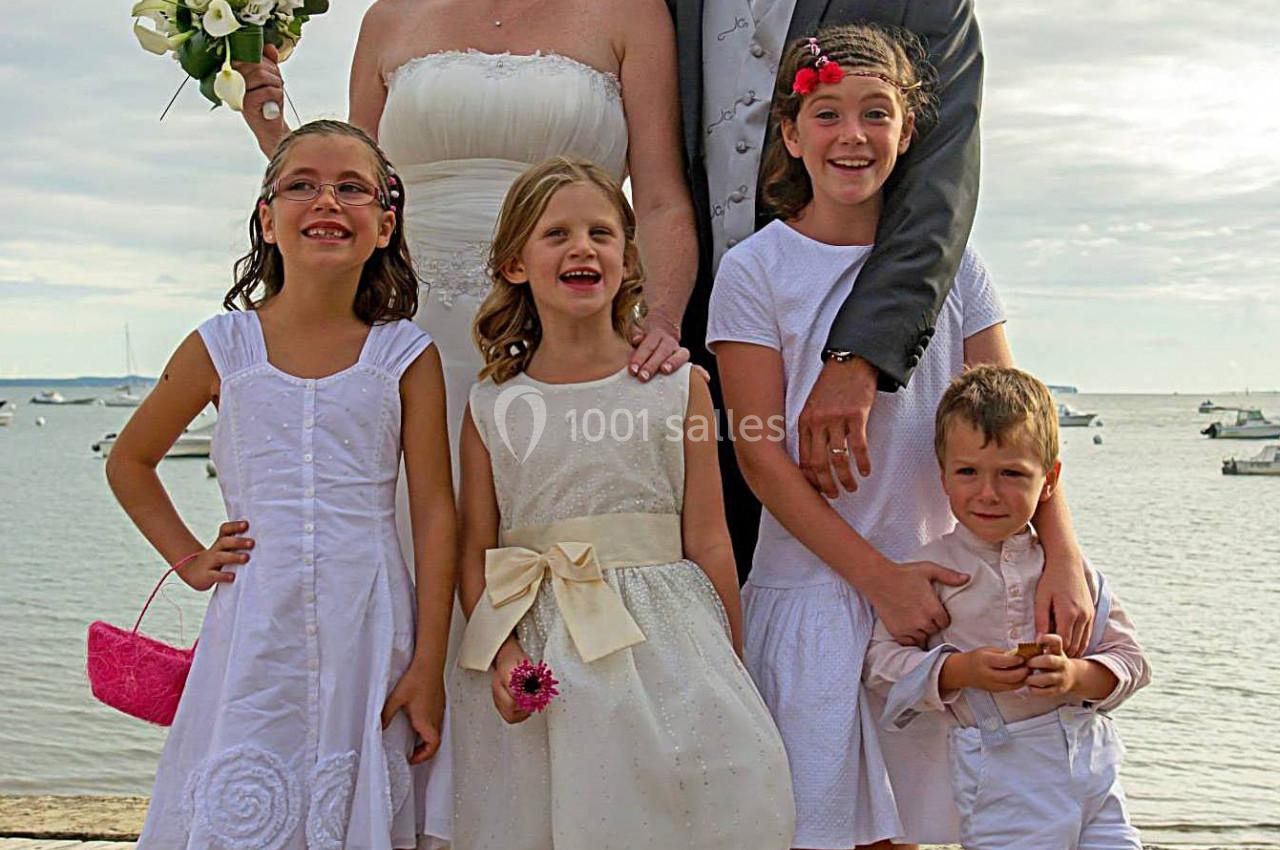Un couple en tenue de mariage pose sur un ponton avec quatre enfants souriants devant un paysage côtier.