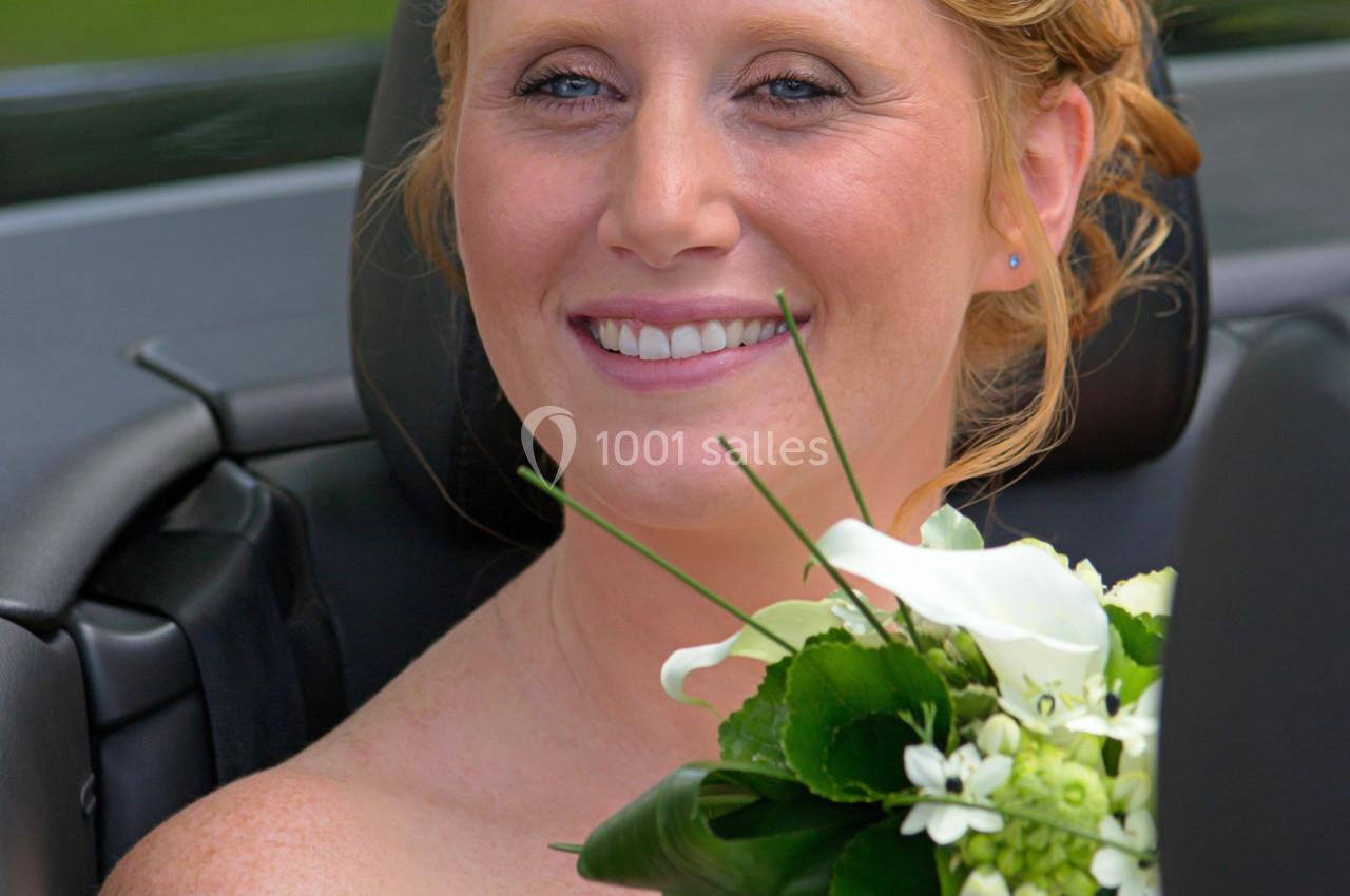 Femme souriante en robe de mariée tenant un bouquet de fleurs blanches, assise dans une voiture décapotable.