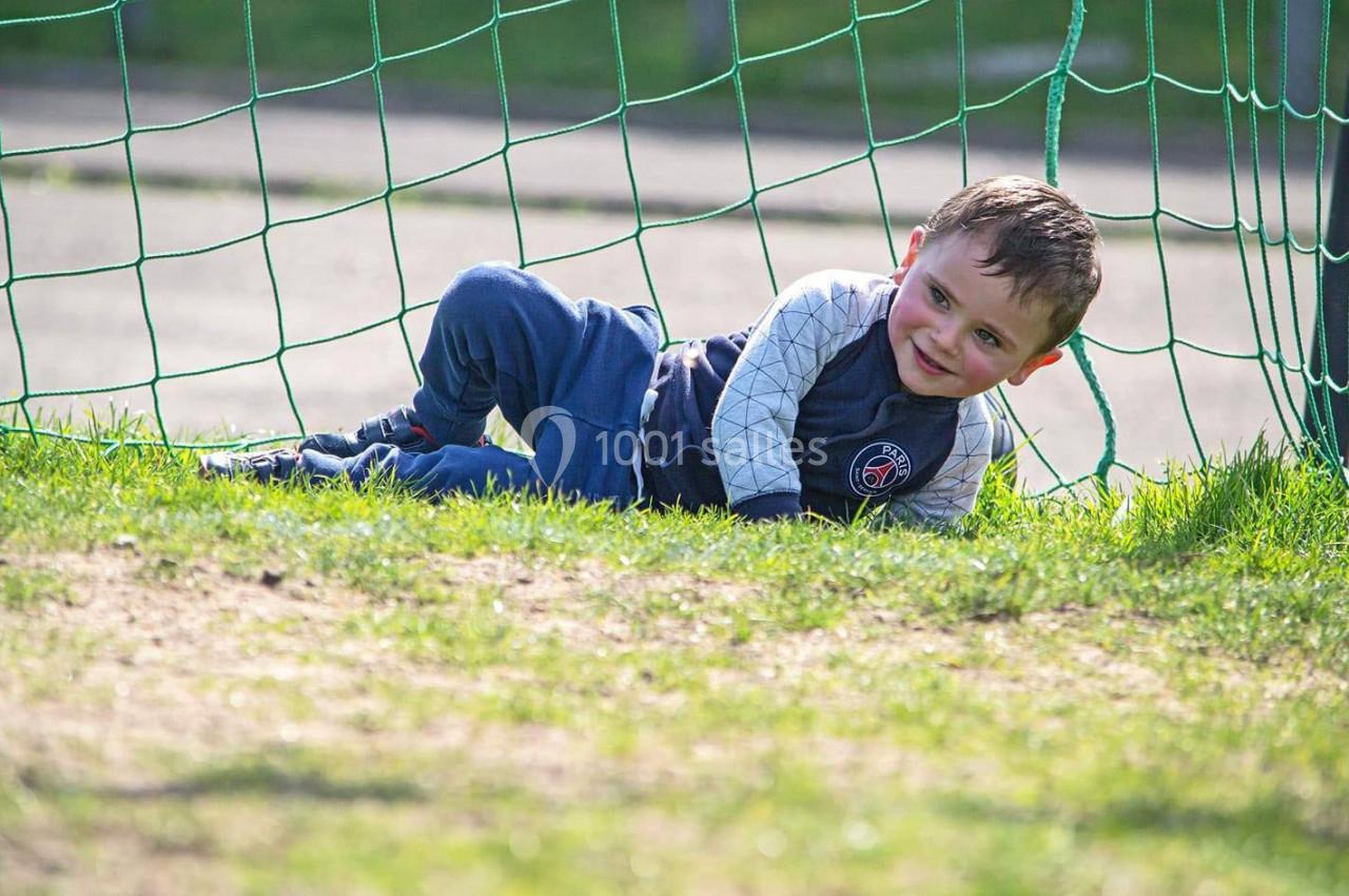 Un enfant souriant allongé sur l'herbe devant un filet de but, dans un espace extérieur ensoleillé.