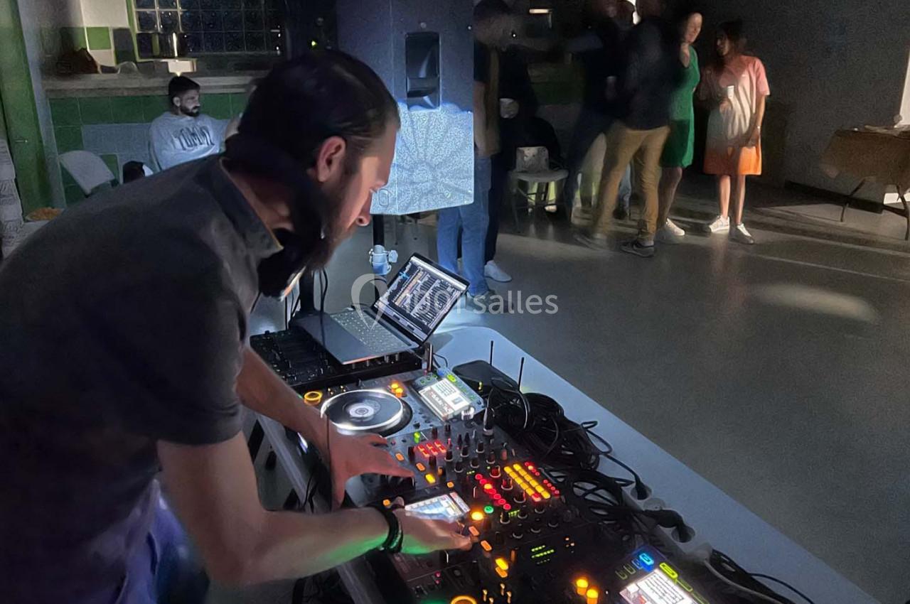 Un DJ mixe de la musique sur une console éclairée, dans une salle avec des personnes en arrière-plan.