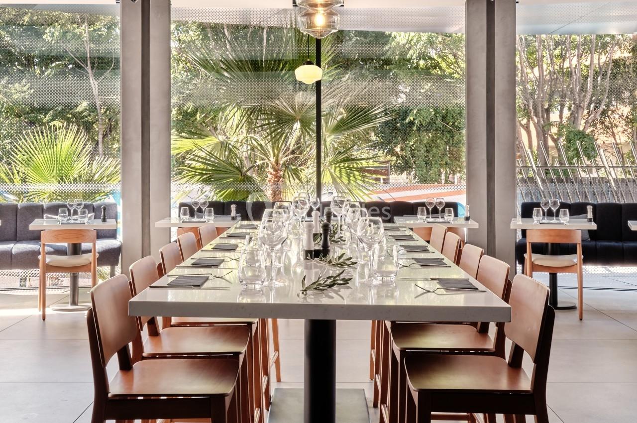 Table rectangulaire avec chaises en bois, dressée avec verres et couverts, dans un espace lumineux avec vue sur des palmiers.