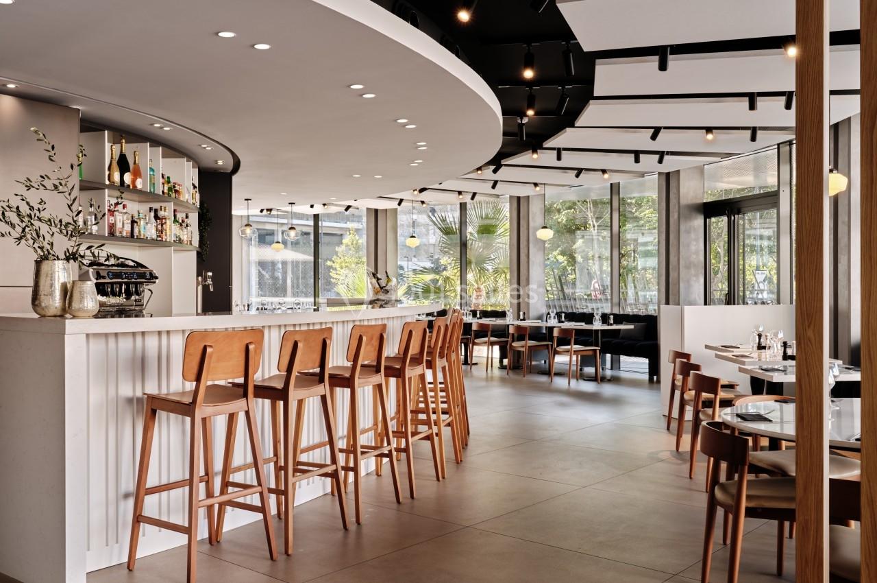 Intérieur d'un restaurant moderne avec un bar, des tabourets en bois et des tables près de grandes baies vitrées.