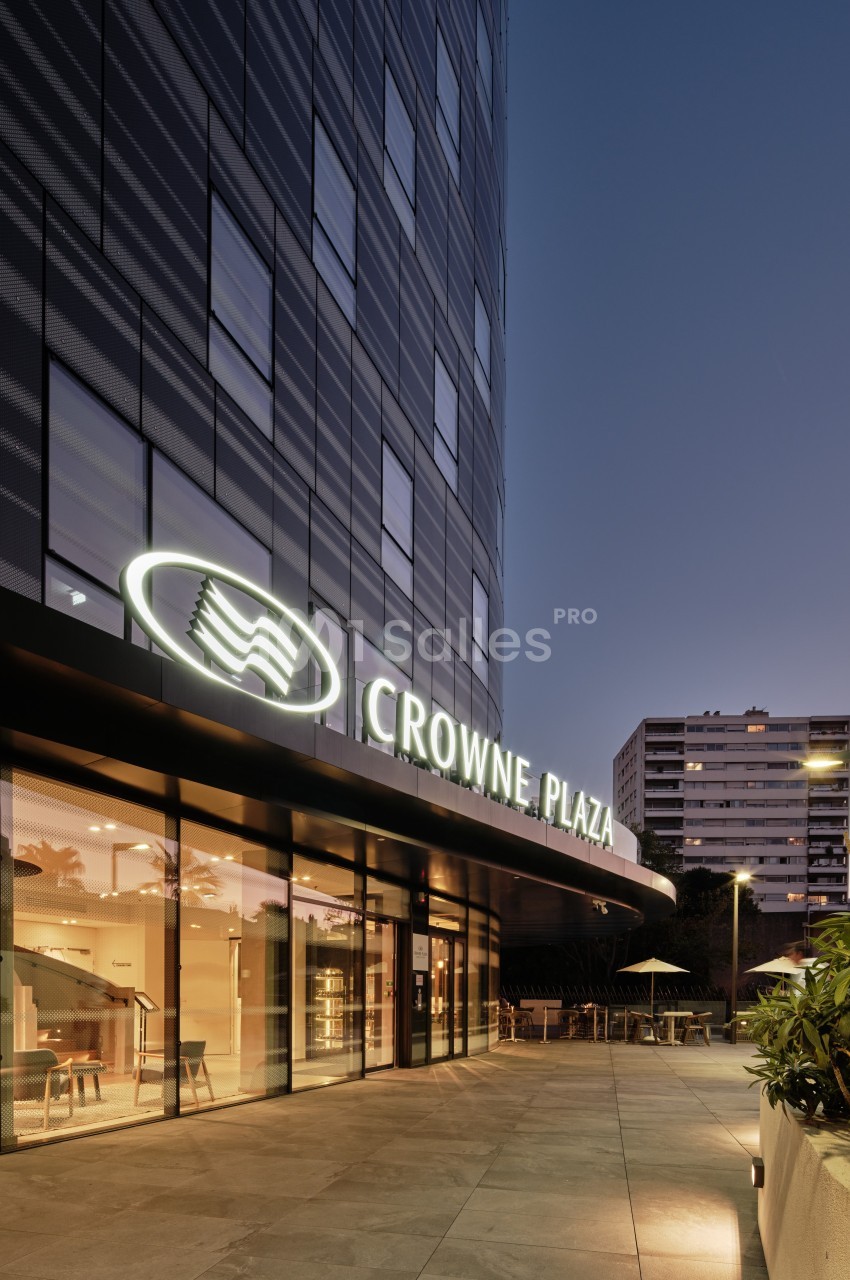Façade d'un hôtel moderne avec enseigne lumineuse ’Crowne Plaza’ au crépuscule, entourée de végétation et d'immeubles.