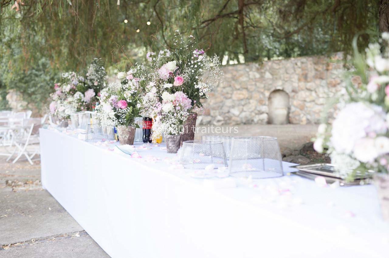 Table décorée avec des bouquets de fleurs roses et blanches, verres et nappes blanches en extérieur près d'un mur en pierre.