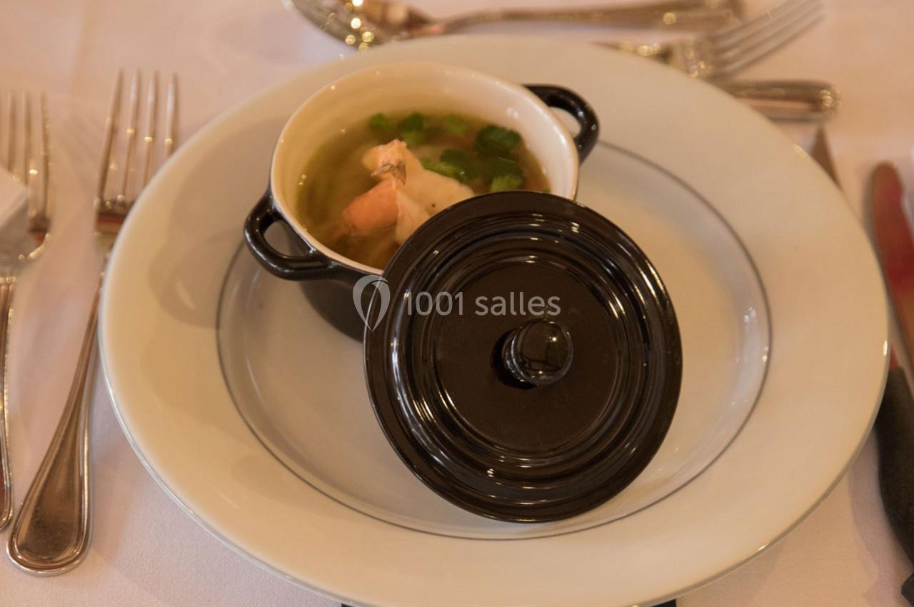 Une petite cocotte noire contenant une soupe avec des légumes et un morceau de viande, posée sur une assiette blanche.