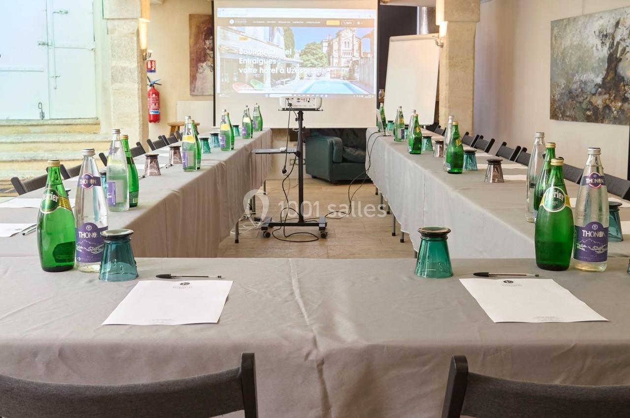 Salle de réunion aménagée en U avec tables couvertes, chaises, bouteilles d'eau, documents et écran de projection.