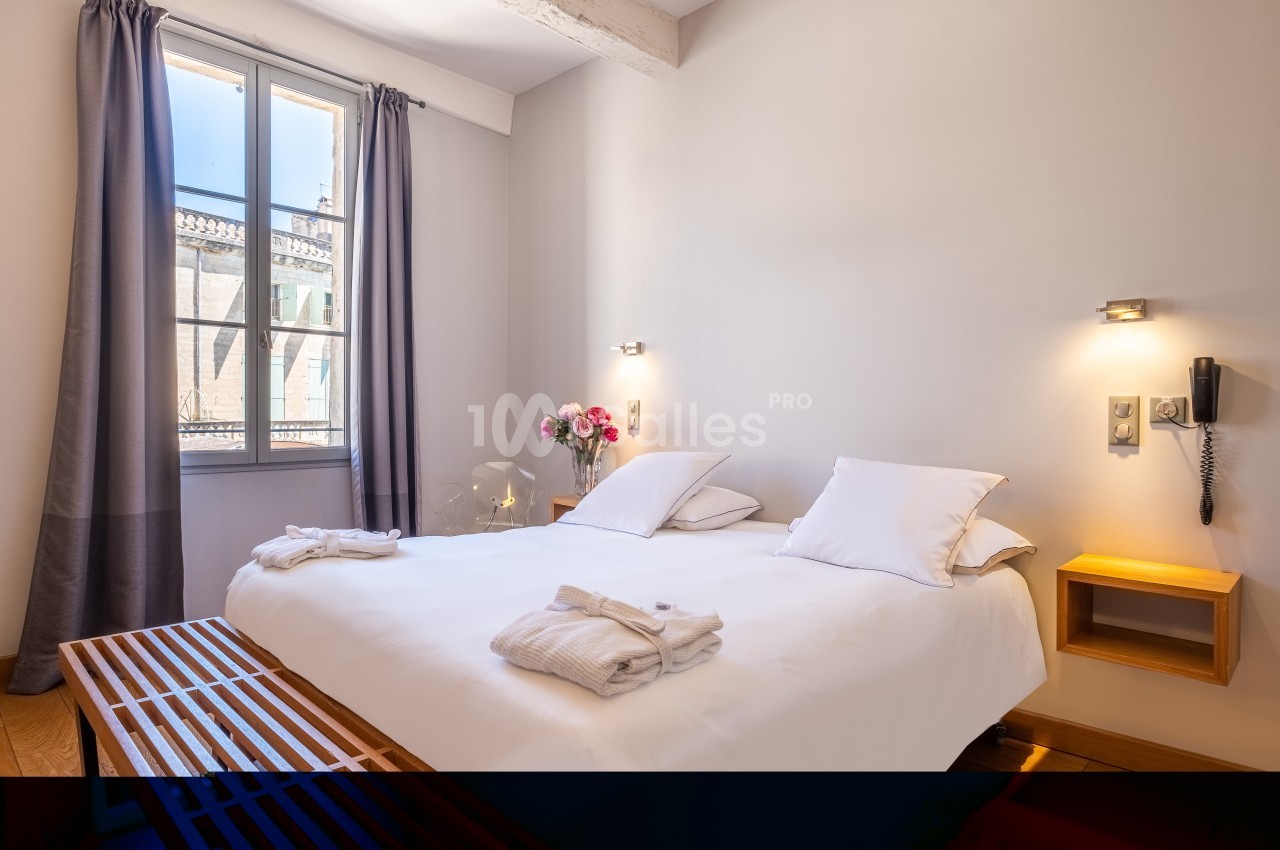 Chambre lumineuse avec un lit double, des draps blancs, des tables de chevet, une fenêtre et des rideaux gris.