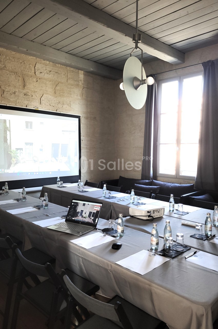 Salle de réunion lumineuse avec table rectangulaire, chaises noires, projecteur, écran et bouteilles d'eau.